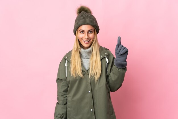 Mujer joven con sombrero de invierno aislado en pared rosa apuntando hacia una gran idea