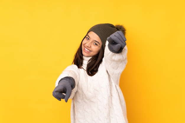 Mujer joven con sombrero de invierno sobre pared amarilla aislada señala con el dedo mientras sonríe