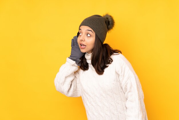 Mujer joven con sombrero de invierno sobre pared amarilla escuchando algo poniendo la mano en la oreja