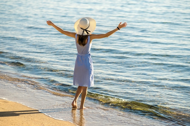 Mujer joven con sombrero de paja y un vestido de pie levantando las manos en la playa de arena vacía en la orilla del mar