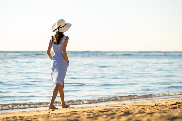 Mujer joven con sombrero de paja y un vestido de pie en la playa vacía