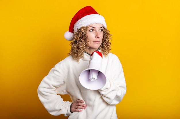 Mujer joven con sombrero de santa claus con un megáfono sobre un fondo amarillo.