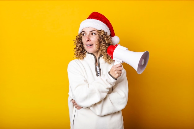 Mujer joven con sombrero de santa claus con un megáfono sobre un fondo amarillo