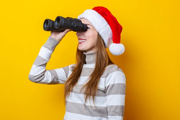 Mujer joven con un sombrero de Santa Claus mira a través de binoculares sobre un fondo amarillo