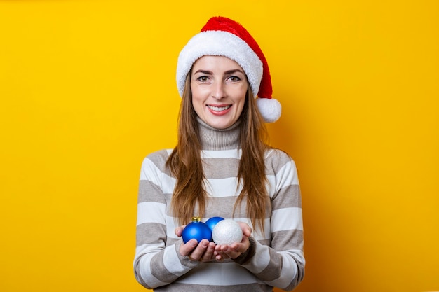 Mujer joven con sombrero de Santa Claus tiene bolas decorativas sobre un fondo amarillo.