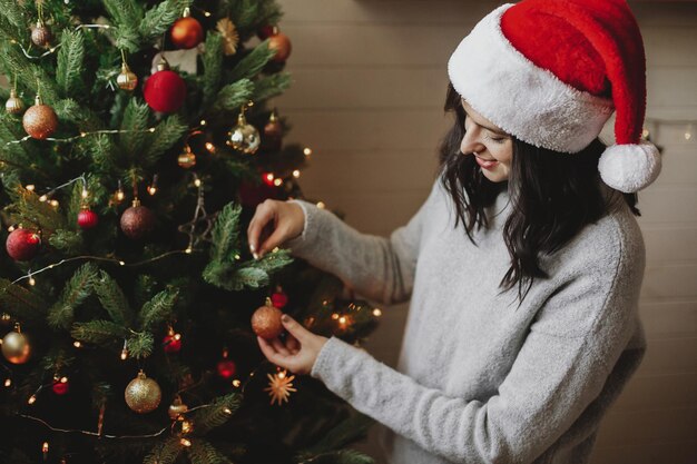 Mujer joven con sombrero de santa decorando el árbol de navidad moderno con adornos de brillo en la sala festiva