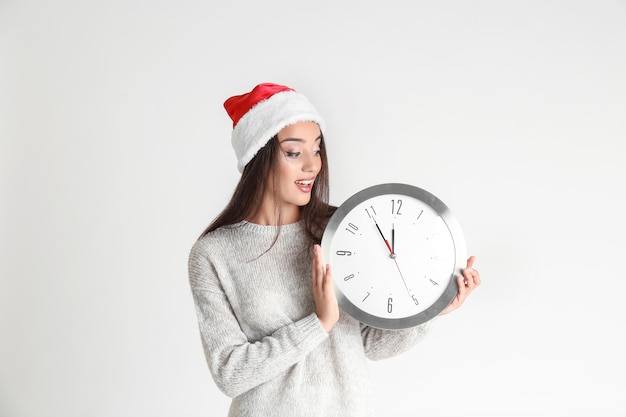 Mujer joven con sombrero de Santa con reloj sobre fondo claro. Concepto de cuenta regresiva de Navidad