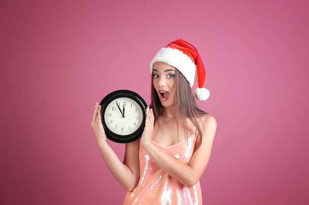 Mujer joven con sombrero de Santa con reloj sobre fondo de color. Concepto de cuenta regresiva de Navidad