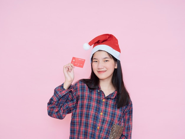 Mujer joven con sombrero de santa con tarjeta de crédito en la pared rosa.