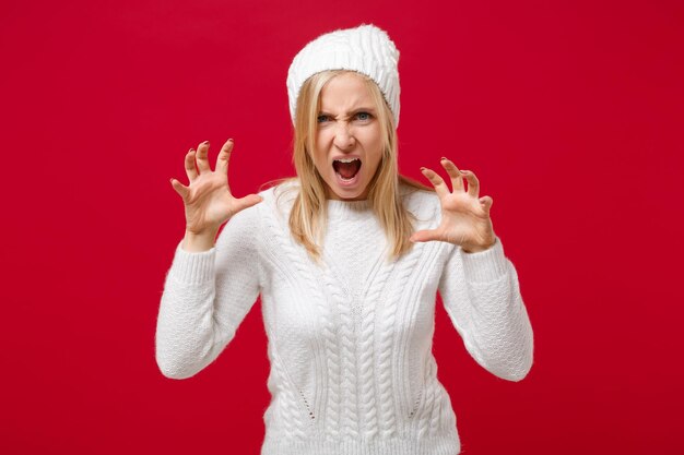 Mujer joven con sombrero de suéter blanco aislado en el fondo de la pared roja en el estudio. Concepto de temporada fría de estilo de vida de moda saludable. Simulacros de espacio de copia. Gritando, gruñendo como un animal, haciendo gestos de garras de gato.
