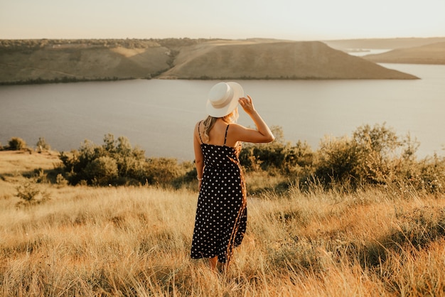 Mujer joven con sombrero en vestido con lunares se encuentra en medio de la pradera en un acantilado sobre el mar al atardecer