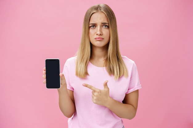 Mujer joven sombría y triste apuntando a la pantalla del teléfono inteligente de pie contra el fondo de color rosa