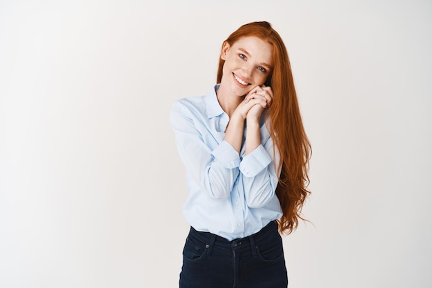 Mujer joven soñadora y romántica con el pelo largo y rojo mirando a la cámara con una sonrisa encantadora admirar o imaginar algo de fondo blanco