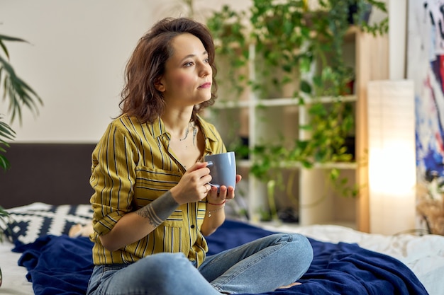 Mujer joven soñadora en ropa casual sentada en la cama en posición de loto bebiendo café o té de hierbas