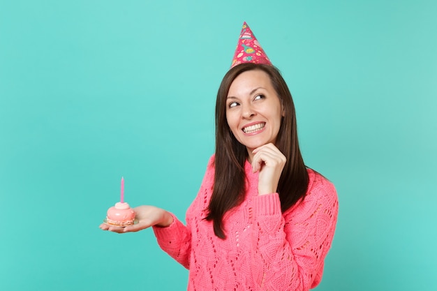 Mujer joven soñadora en suéter rosa de punto, sombrero de cumpleaños mirando hacia arriba, coloque el apoyo de la mano en la barbilla, sostenga la torta en la mano con vela aislada sobre fondo azul. Concepto de estilo de vida de personas. Simulacros de espacio de copia.