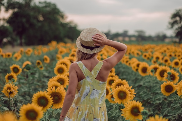 Mujer joven soñando en vestido amarillo sosteniendo un sombrero con una mano y alejándose en un campo de girasoles en verano, vista desde su espalda. Viendo hacia adelante. copia espacio