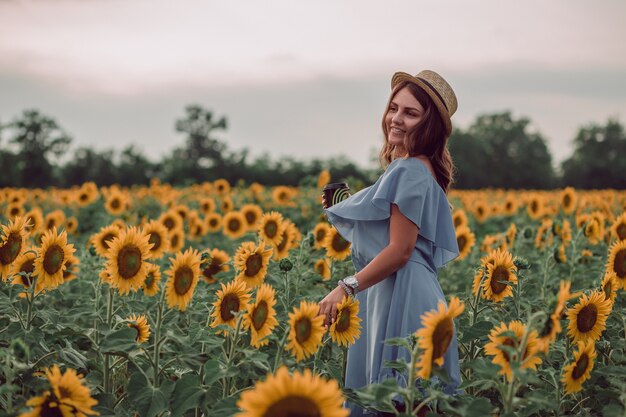 Mujer joven soñando con vestido azul y sombrero sosteniendo una taza de café en un campo de girasoles en verano, vista desde su lado. Mirando hacia un lado. Sonriente. copia espacio