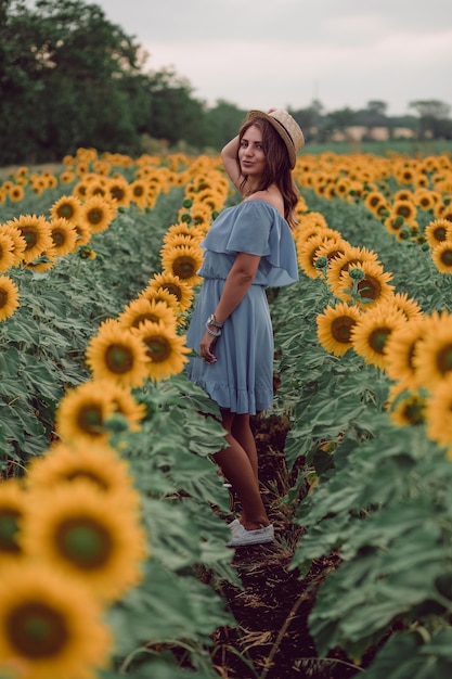 Mujer joven soñando en vestido azul sosteniendo un sombrero en un campo de girasoles en verano, vista desde su lado. Mirando hacia un lado. Cuerpo completo. copia espacio