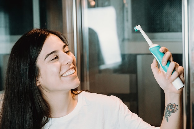 Mujer joven sonriendo a un cepillo de dientes eléctrico en el baño