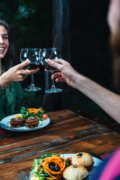 Mujer joven sonriendo y disfrutando de una cena vegetariana romántica servida en un restaurante brindando con vino rojo en una cita sentada en una mesa de madera con un traje verde colorido