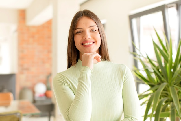 Mujer joven sonriendo disfrutando de la vida sintiéndose feliz amistoso satisfecho y despreocupado con la mano en la barbilla