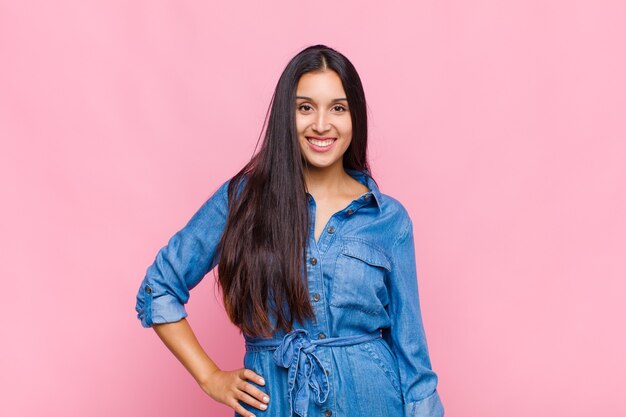 Mujer joven sonriendo felizmente con una mano en la cadera y actitud confiada, positiva, orgullosa y amistosa