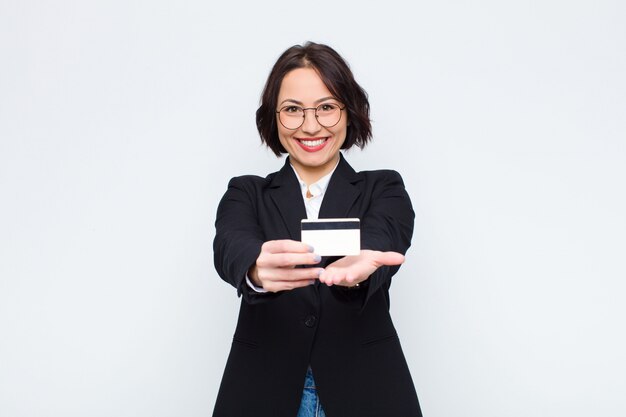 Mujer joven sonriendo felizmente con mirada amigable, segura y positiva, ofreciendo y mostrando un objeto o concepto con una tarjeta de crédito