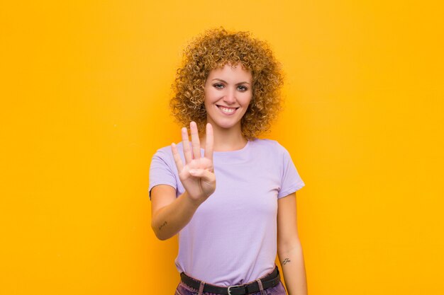 Mujer joven sonriendo y mirando amigable, mostrando el número cuatro o cuarto con la mano hacia adelante, contando hacia atrás sobre la pared naranja