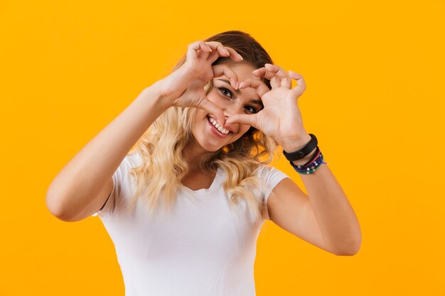Mujer joven sonriendo y mostrando en forma de corazón con los dedos, aislado sobre pared amarilla