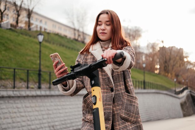 Mujer joven sonriente con abrigo que usa un teléfono inteligente para alquilar scooter eléctrico en la calle de la ciudad de otoño Vehículo de transporte ecológico Tecnologías modernas Actividades al aire libre