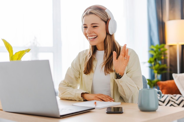 Mujer joven sonriente con auriculares agitando durante una llamada de video en la oficina en casa