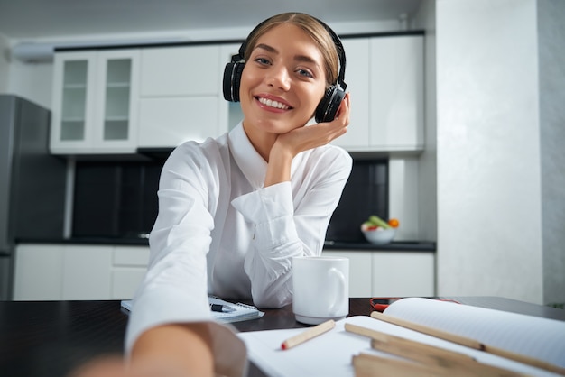 Mujer joven sonriente en auriculares inalámbricos con videollamada en la computadora portátil mientras trabaja en casa