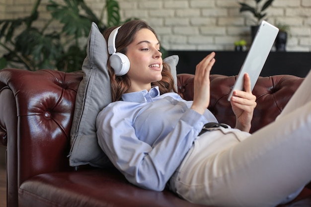 Mujer joven sonriente con auriculares y tableta digital en el sofá.