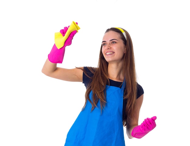 Mujer joven sonriente en camiseta azul y delantal con guantes rosas sosteniendo cosas de limpieza en un lavabo