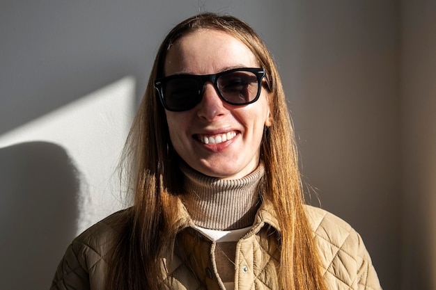 Mujer joven sonriente en una chaqueta con gafas de sol contra una pared blanca