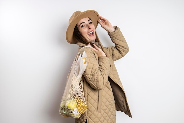 Mujer joven sonriente con una chaqueta en un sombrero con una bolsa de compras contra una pared blanca