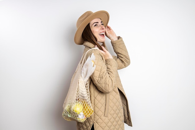 Mujer joven sonriente con una chaqueta en un sombrero con una bolsa de compras contra una pared blanca