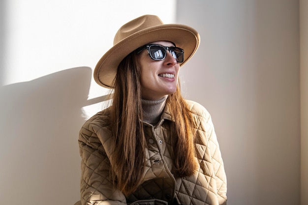 Mujer joven sonriente con una chaqueta en un sombrero con gafas de sol contra una pared blanca