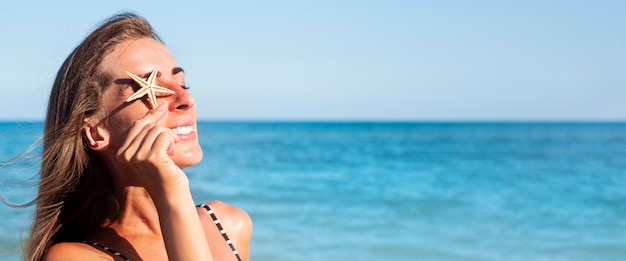 La mujer joven sonriente cierra su estrella de mar del ojo en la playa. Bandera.