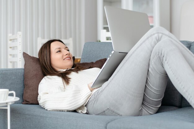 Mujer joven sonriente con una computadora portátil tirada en el sofá en la sala de estar