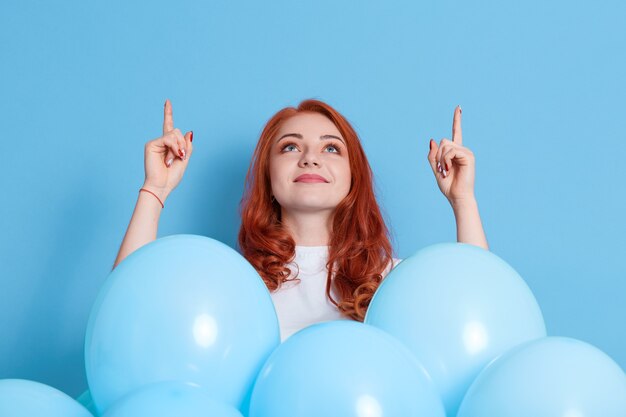 Mujer joven sonriente en cuello alto blanco apuntando los dedos índices hacia arriba con ambas manos, celebrando y sosteniendo globos de aire azul aislados en la pared de color. Fiesta de cumpleaños.