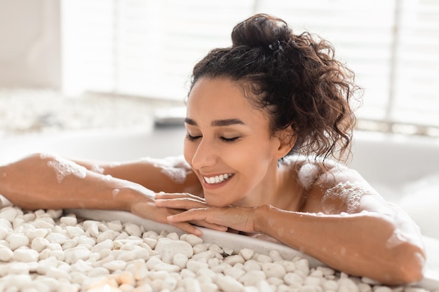 Mujer joven sonriente descansando en la bañera con los ojos cerrados disfrutando de un baño caliente con espuma en casa