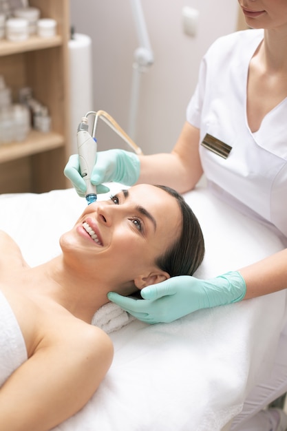 Mujer joven sonriente disfrutando del proceso de nutrición de la piel en la clínica moderna con la ayuda de una herramienta especial