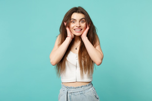 Mujer joven sonriente emocionada en ropa casual ligera que mira la cámara, poniendo las manos en la cara aislada en el fondo azul turquesa de la pared. Personas sinceras emociones, concepto de estilo de vida. Simulacros de espacio de copia.