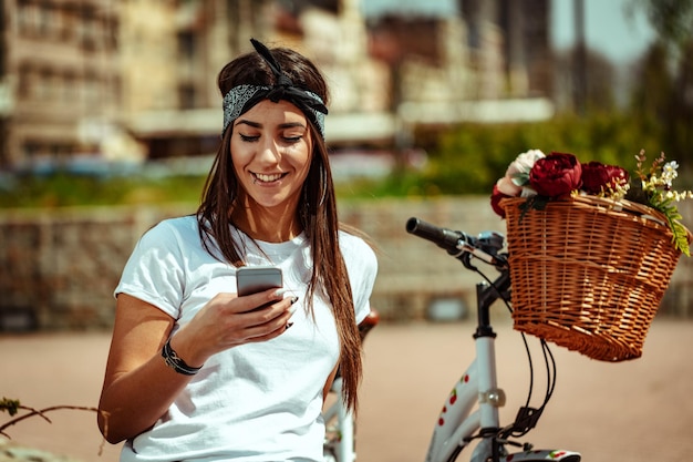 Mujer joven sonriente enviando mensajes de texto en un teléfono inteligente en la calle de la ciudad, en un día soleado, al lado de la bicicleta con una cesta de flores.