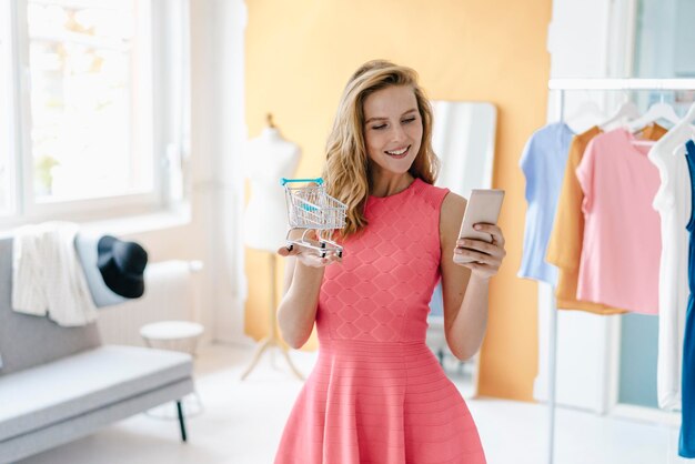 Mujer joven sonriente en el estudio de moda tomando una foto de teléfono celular del modelo de carrito de compras