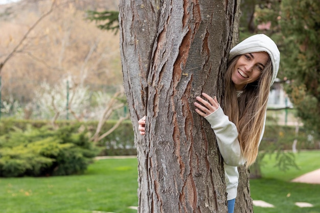 Mujer joven sonriente y feliz detrás del tronco del árbol con una capucha de sudadera