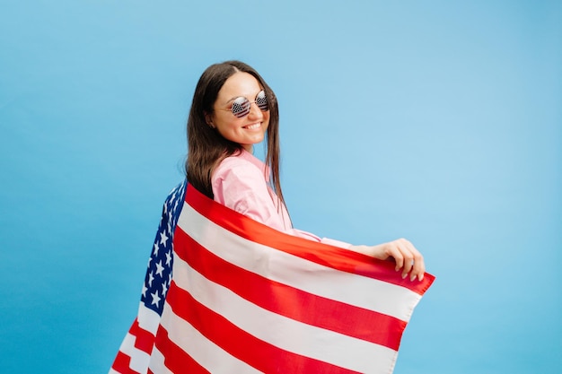 Mujer joven sonriente feliz lleva ropa casual sosteniendo bandera estadounidense y mirando a la cámara aislada en fondo de color azul