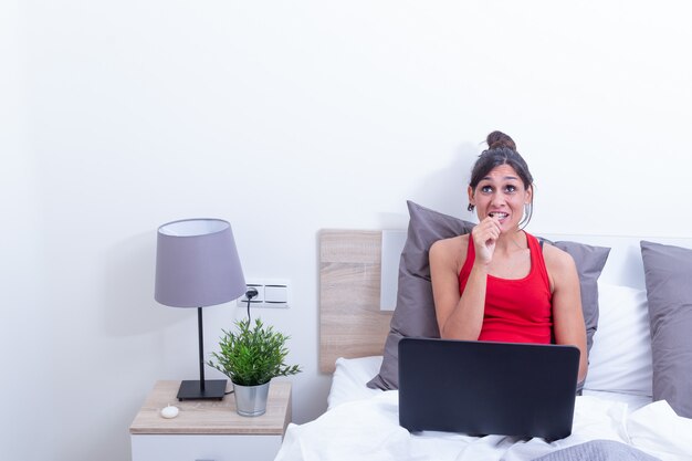 Mujer joven sonriente feliz, relajándose en su cama con la computadora portátil. Mira pensativo.