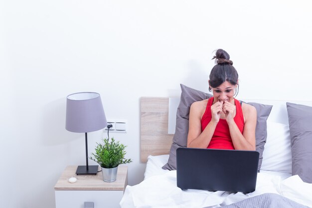 Mujer joven sonriente feliz, relajándose en su cama con la computadora portátil. Se ve nervioso en la computadora portátil.